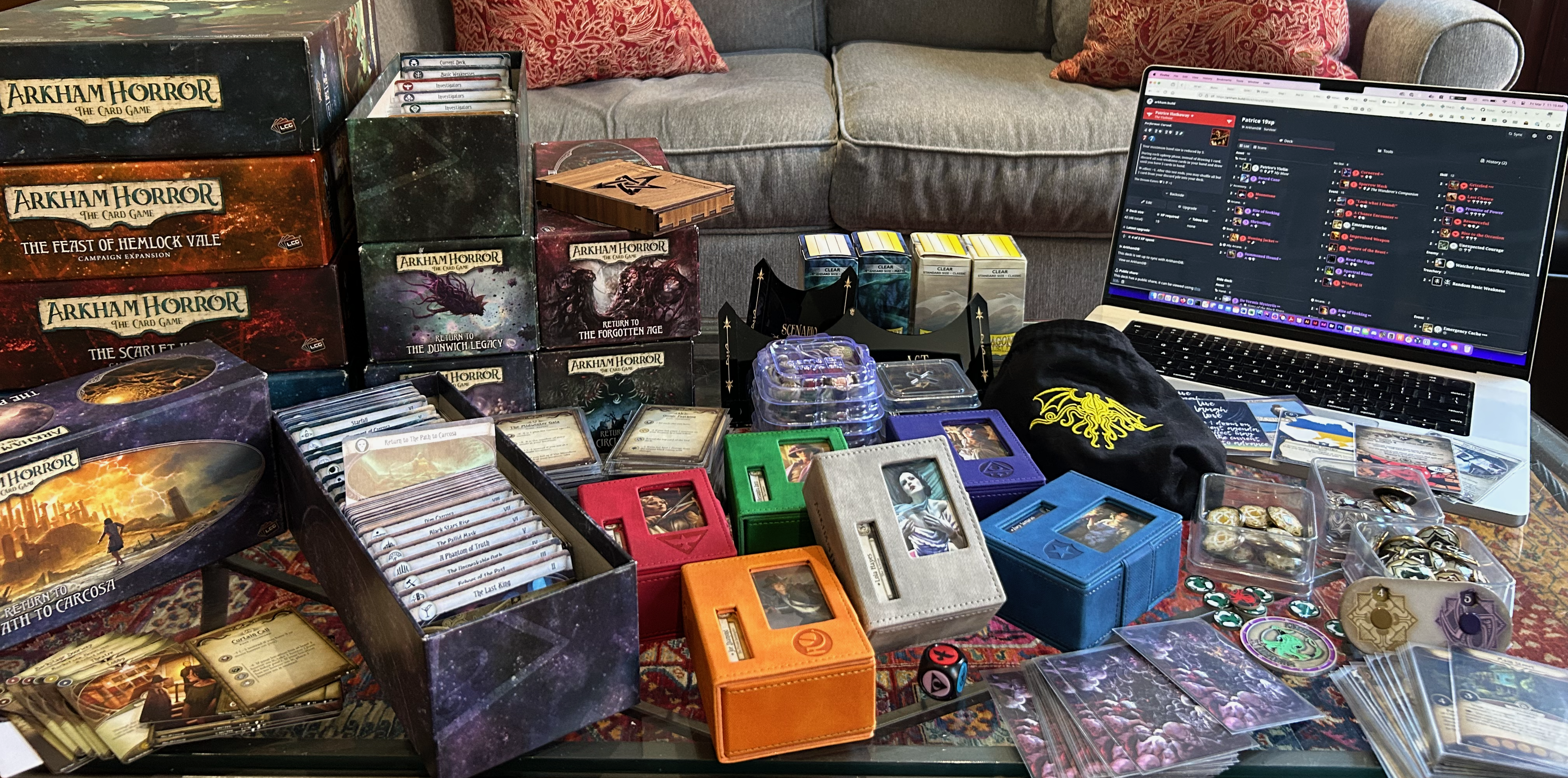 A coffee table laden with AHLCG boxes, cabochon tokens, scenarios, promo cards from previous cons, AH deck boxes, sleeves, chaos bag, and a laptop open to an arkham.build screen