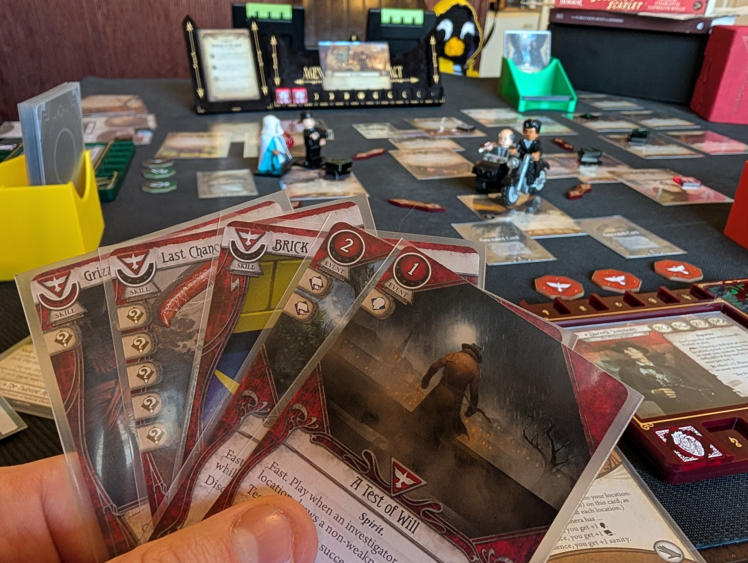 Closeup of a fan of Arkham Horror cards in a player's hand, with the game set up in the background